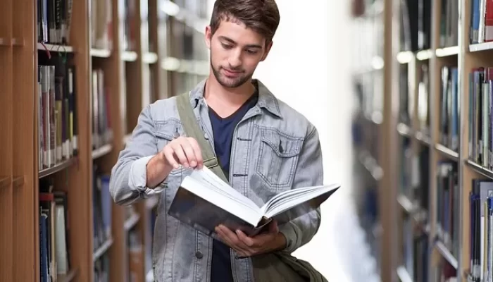 Ein junger Mann blättert in einem Buch und steht zwischen den Regalen einer Bibliothek, nachdem er mit A2B Umzüge in sein neues Zuhause gezogen ist.